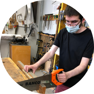 Zane standing at a wooden bench using an orange handled saw to cut through a peice of wood that is in a vice
